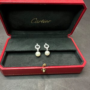 May include: A red Cartier jewelry box with a black interior. Two diamond and pearl earrings are displayed inside the box.