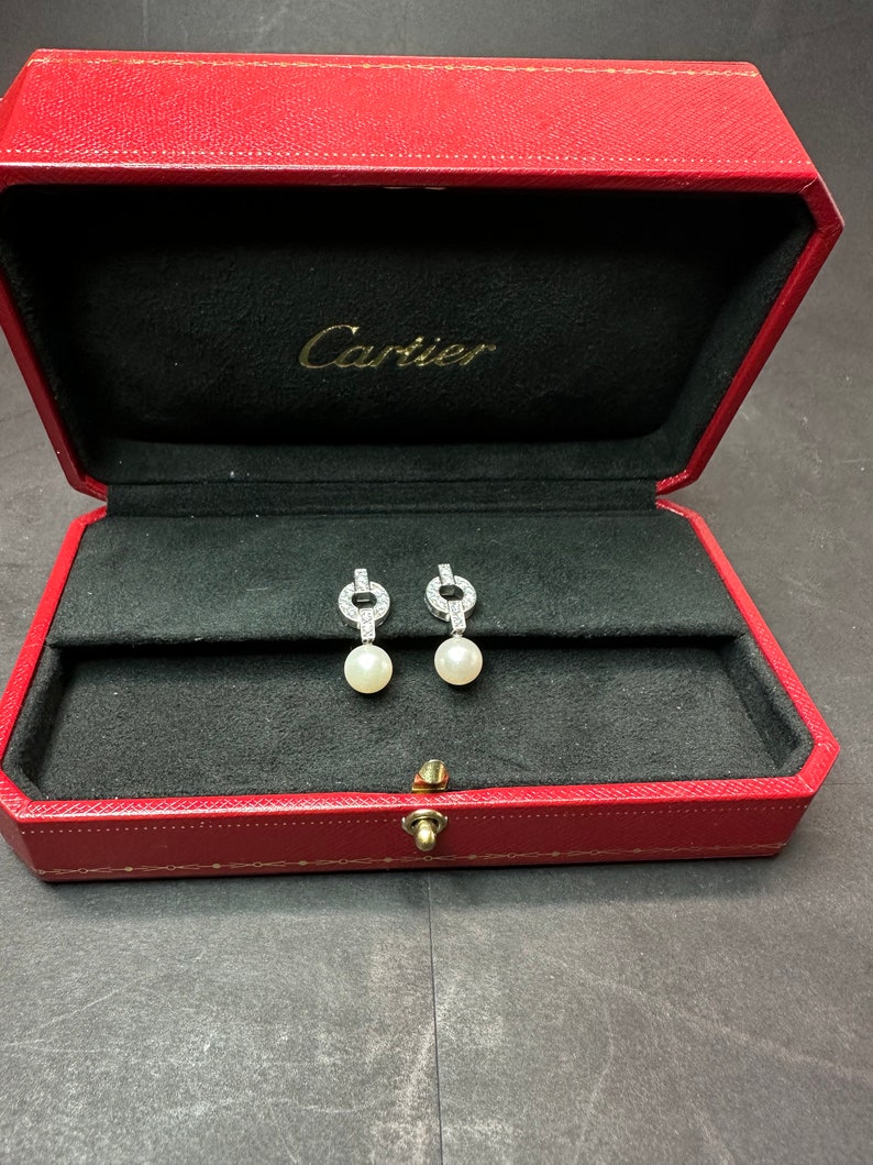 May include: A red Cartier jewelry box with a black interior. Two diamond and pearl earrings are displayed inside the box.