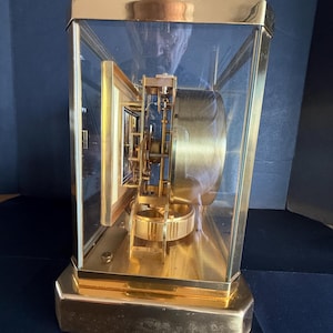 May include: A gold-toned, rectangular clock with a glass enclosure. The clock's face is visible through the glass, showcasing the intricate internal mechanisms. The clock sits on a gold base and has a gold frame.