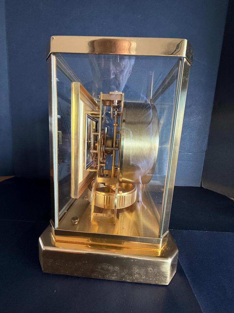 May include: A gold-toned, rectangular clock with a glass enclosure. The clock's face is visible through the glass, showcasing the intricate internal mechanisms. The clock sits on a gold base and has a gold frame.