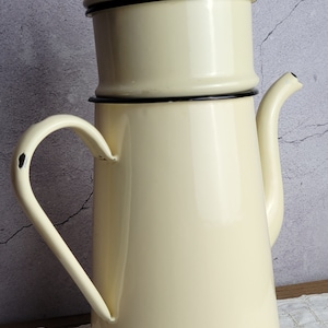 May include: A cream-colored enamel coffee pot with a black rim and a lid. The pot has a spout and a handle.