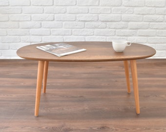 Puede incluir: Una mesa de centro ovalada de madera marrón con acabado de veta de madera y patas de madera cónicas. Una taza de cerámica blanca y una revista están sobre la mesa.