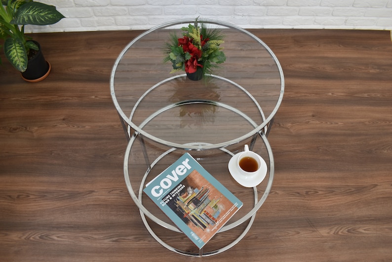 Puede incluir: Una mesa de centro de vidrio de dos niveles con un marco cromado. El nivel superior tiene un ramo de flores rojas en una maceta negra. El nivel inferior tiene una revista y una taza de t&eacute; en un platillo.