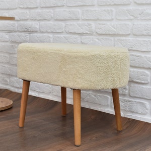 May include: A white fluffy ottoman with four wooden legs. The ottoman is sitting on a wooden floor in front of a white brick wall.