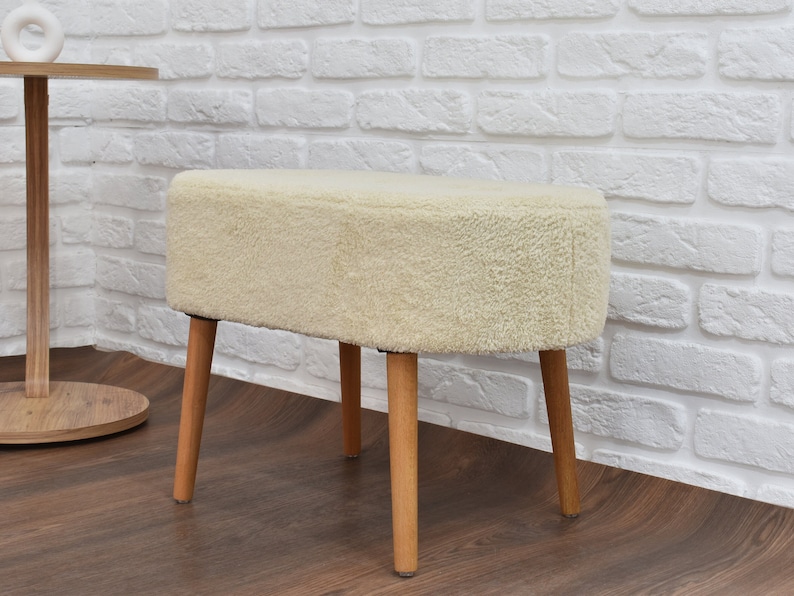 May include: A white fluffy ottoman with four wooden legs. The ottoman is sitting on a wooden floor in front of a white brick wall.