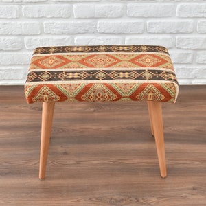 Puede incluir: Un pequeño taburete de madera con un asiento de tela estampado de colores. La tela presenta un diseño geométrico en tonos rojos, naranjas, amarillos, verdes y negros.