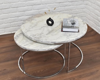 May include: A set of two round nesting tables with a white marble top and chrome legs. The top table has a book on it and a small black vase with dried flowers.