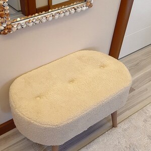 May include: A white oval-shaped ottoman with a fluffy, plush texture and three wooden legs. The ottoman is sitting on a light wood floor.