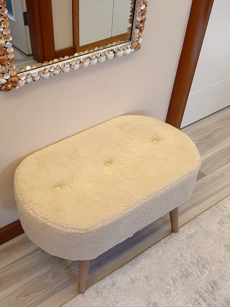 May include: A white oval-shaped ottoman with a fluffy, plush texture and three wooden legs. The ottoman is sitting on a light wood floor.
