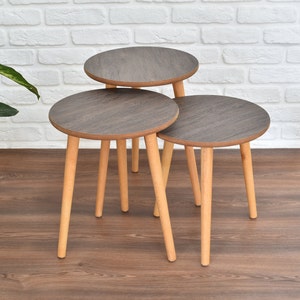 Puede incluir: Tres mesas nido redondas de madera con acabado en madera de grano marrón oscuro y patas de madera marrón claro.
