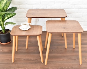 May include: Three wooden nesting tables with a light brown wood grain finish and natural wood legs. The tables are arranged in a staggered formation, with the largest table at the back and the smallest table in the front. A white coffee cup and saucer are sitting on the top of the smallest table.