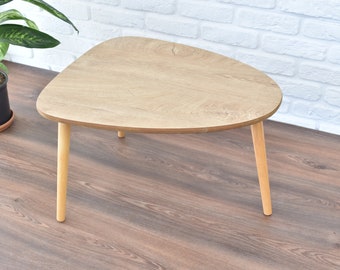 Puede incluir: Una mesa de centro triangular de madera clara con un borde redondeado y tres patas de madera.