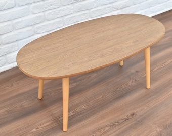 May include: A light brown oval-shaped coffee table with three wooden legs.