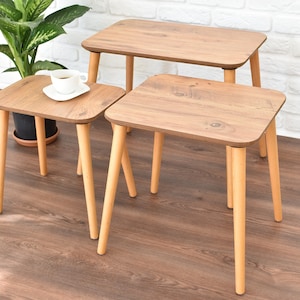 Puede incluir: Tres mesas nido de madera con un acabado en madera de grano marrón claro y patas de madera natural. Las mesas están dispuestas en forma escalonada, con la mesa más grande en la parte posterior y la mesa más pequeña en la parte delantera.