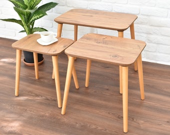 May include: Three wooden nesting tables with a light brown wood grain finish and natural wood legs. The tables are arranged in a staggered formation, with the largest table in the back and the smallest table in the front.