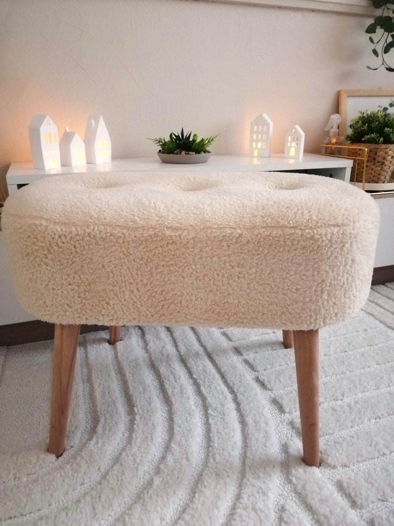 May include: A white oval-shaped ottoman with a fluffy, cream-colored fabric cover. The ottoman has four wooden legs and is sitting on a white shag rug.
