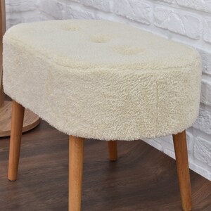 May include: A white fluffy oval-shaped ottoman with four wooden legs. The ottoman has a tufted top with three buttons.
