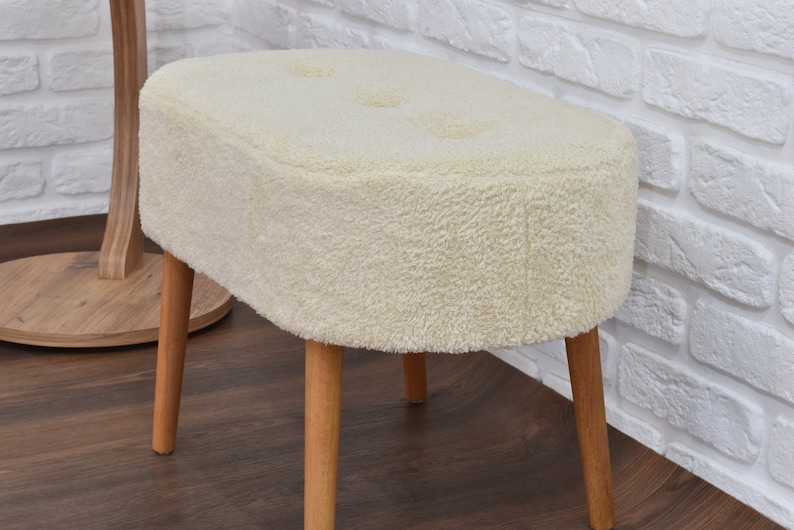 May include: A white fluffy oval-shaped ottoman with four wooden legs. The ottoman has a tufted top with three buttons.