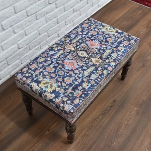 May include: A wooden ottoman with a dark blue and floral patterned fabric top. The ottoman has four legs and is sitting on a wooden floor.