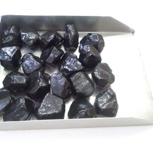 May include: A tray of black gemstones, likely raw, unpolished, and rough cut. The stones are various sizes and shapes, with a glossy, dark finish.