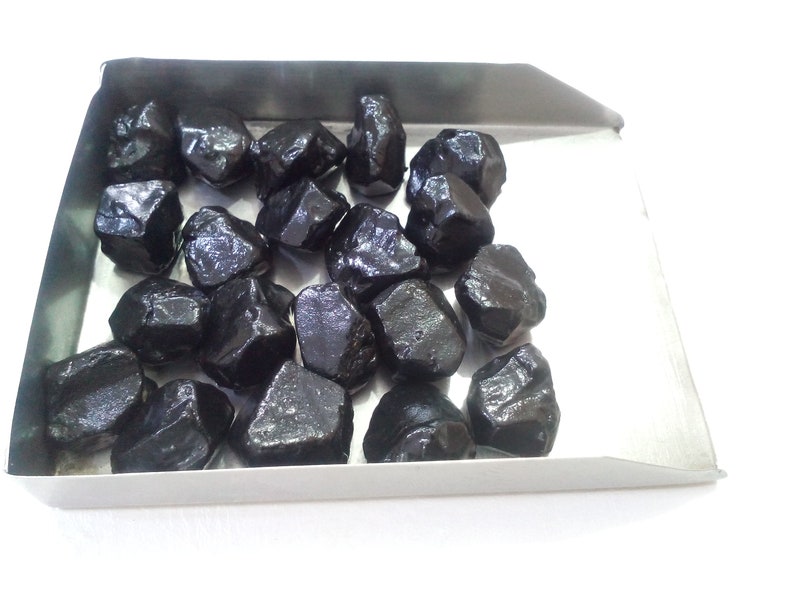 May include: A tray of black gemstones, likely raw, unpolished, and rough cut. The stones are various sizes and shapes, with a glossy, dark finish.