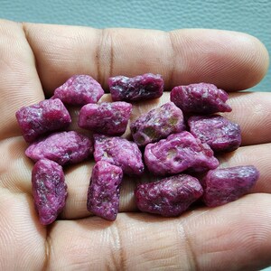 Può includere: Una collezione di gemme di rubino grezze e non tagliate. I rubini grezzi mostrano una tonalità rosso-rosa intensa, con una superficie ruvida e irregolare. Le gemme variano per dimensioni e forma, mostrando il loro stato naturale.