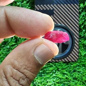 Puede incluir: Una pequeña piedra preciosa en bruto de color rosa vibrante con una superficie irregular y texturizada. La piedra es translúcida, permitiendo que la luz atraviese su estructura cristalina, revelando tonos magenta profundos y facetas naturales.