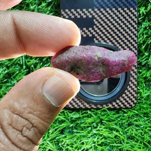 Puede incluir: Una piedra preciosa de rubí en bruto y sin pulir con una superficie rugosa y texturizada y una coloración magenta rosada profunda. La piedra muestra inclusiones minerales naturales y una forma irregular y alargada, revelando su opacidad.