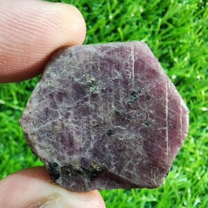 May include: A raw, hexagonal-shaped ruby crystal with a deep purplish-red hue. The surface of the gemstone displays a natural, textured appearance with visible inclusions, dark mineral flecks, and subtle crystalline striations across its face.