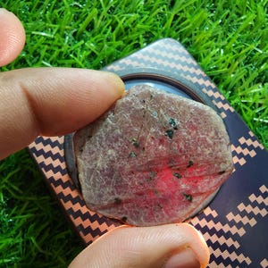 May include: A rough, hexagonal-shaped ruby gemstone with a mottled pinkish-red surface featuring small dark inclusions. The stone is translucent, showing a bright red glow when backlit, and is positioned against a dark, carbon-fibre patterned surface.