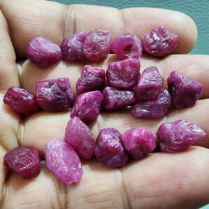 Puede incluir: Una colección de gemas de rubí en bruto sin cortar. Las piedras varían en tamaño y forma, mostrando un tono rojo-rosa intenso. La textura rugosa y la forma natural de los rubíes son claramente visibles, mostrando su belleza sin refinar.