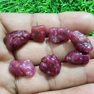 Può includere: Una collezione di gemme di rubino grezze e non tagliate. Le gemme sono di un colore rosso intenso e ricco con un aspetto leggermente traslucido. I rubini sono tenuti in una mano aperta su uno sfondo verde.