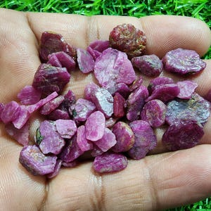 Peut inclure: Un ensemble de pierres précieuses de rubis brutes, non taillées, de formes et de tailles variées. Les rubis présentent des nuances de rose et de rouge profond, avec une surface rugueuse et texturée. Les pierres sont tenues dans une main ouverte.