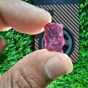 May include: A small, raw, deep pink ruby gemstone with a textured, irregular surface. The stone is translucent, allowing light to pass through its crystalline structure, revealing natural inclusions and a faceted, unpolished appearance.