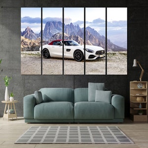 May include: A white convertible sports car with a red interior is parked on a dirt road in front of a mountain range. The car has a black roof and black wheels. The mountains are in the background and are covered in snow.