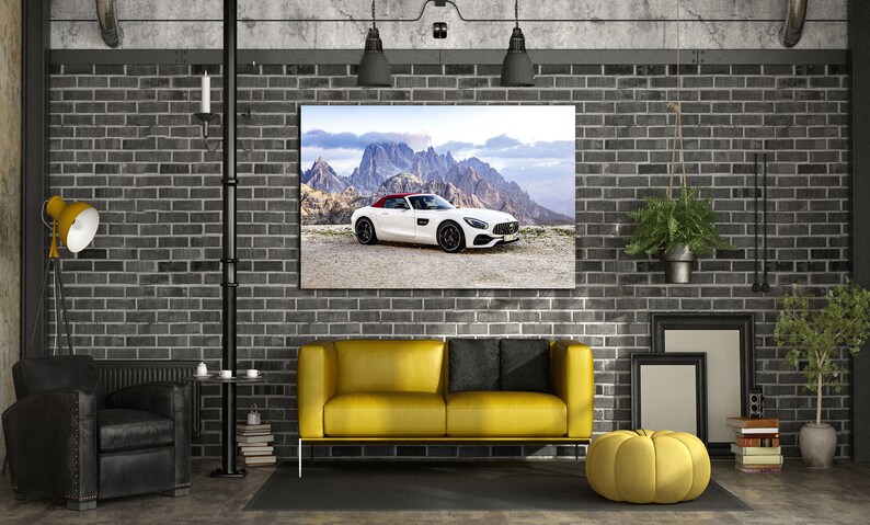 May include: A white convertible sports car with a red roof is featured in a framed print hanging on a brick wall. The print is above a yellow sofa with black pillows. The room is decorated in a modern industrial style with exposed brick and metal accents.