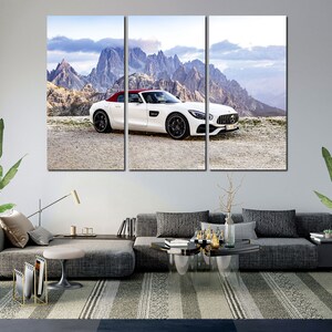 May include: A white Mercedes-Benz AMG GT Roadster with a red convertible top is parked on a gravel road in front of a mountain range. The car is facing the camera and the mountains are in the background.