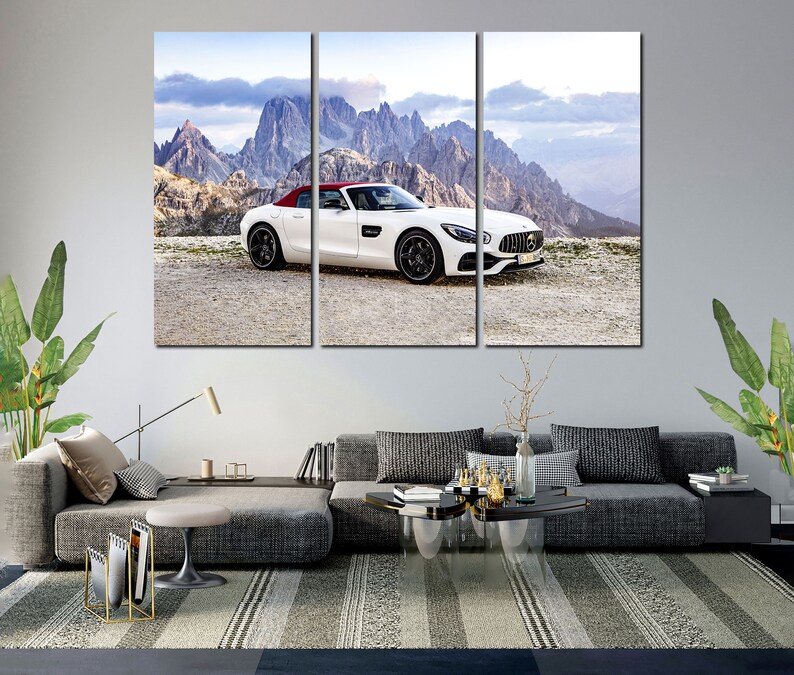May include: A white Mercedes-Benz AMG GT Roadster with a red convertible top is parked on a gravel road in front of a mountain range. The car is facing the camera and the mountains are in the background.