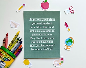 May include: A green card with white text that reads "May the Lord bless you and protect you. May the Lord smile on you and be gracious to you. May the Lord show you his favor and give you his peace." Numbers 6:24-26. The card is surrounded by colorful school supplies, including crayons, a pencil, a paper airplane, a lightbulb, a paintbrush, a pair of scissors, a paint palette, and a globe.