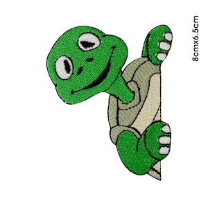 May include: Green and beige embroidered iron-on patch of a cartoon turtle peeking around a corner. The turtle is smiling and has large eyes. The patch measures 8 cm by 6.5 cm.
