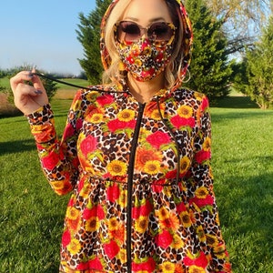 Puede incluir: Una sudadera con capucha negra con cremallera con estampado de leopardo y un patrón de girasoles y rosas. La sudadera tiene una cintura ajustada y una capucha con cordón. La modelo lleva una máscara facial con tachuelas de diamantes de imitación.