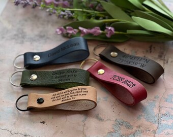 May include: A collection of leather keychains in various colors, including navy, green, brown, and red. Each keychain features engraved text, such as names, coordinates, and inspirational quotes. The keychains have metal key rings and decorative studs.