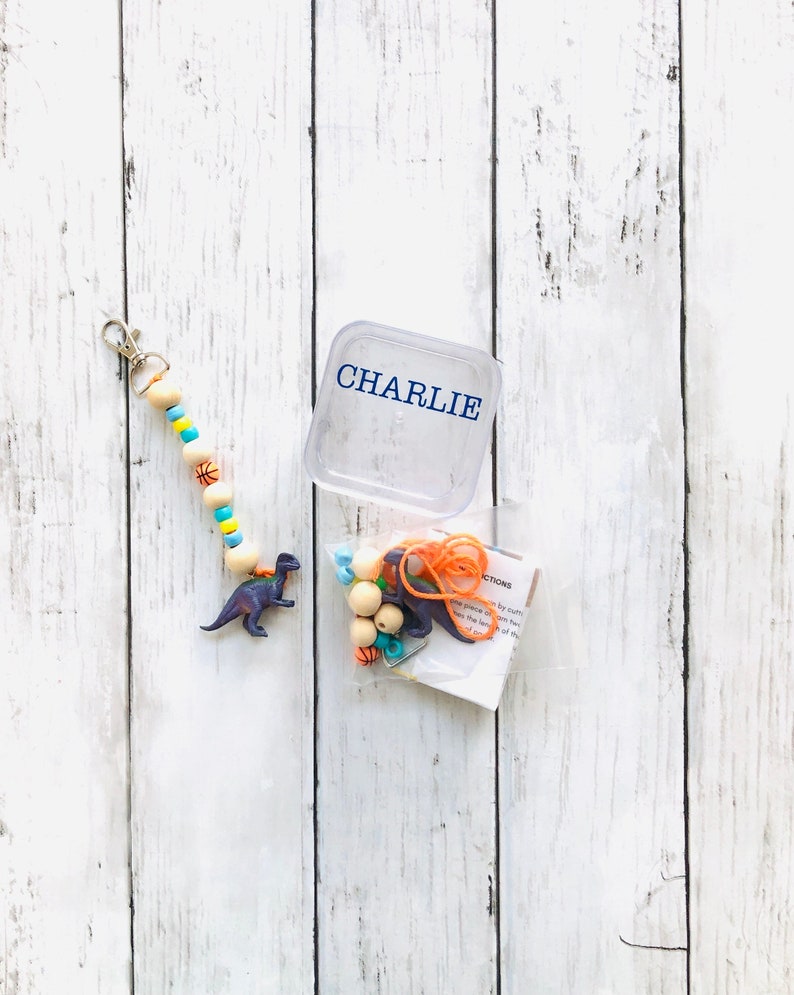 K&ouml;nnte beinhalten: Eine durchsichtige Plastikbox mit dem Namen "CHARLIE" darauf gedruckt. Eine Plastikt&uuml;te enth&auml;lt eine Schnur aus Holzperlen mit orangefarbenen, blauen und gelben Akzenten, eine kleine Dinosaurierfigur und ein St&uuml;ck orangefarbenes Garn.