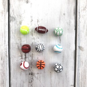 K&ouml;nnte beinhalten: Neun Miniatur-Sportb&auml;lle, darunter ein Tennisball, ein American Football, ein Fu&szlig;ball, ein Basketball, ein Volleyball, ein Baseball und zwei weitere Fu&szlig;b&auml;lle. Die B&auml;lle sind wei&szlig; mit verschiedenen farbigen Details und in drei Reihen auf einer wei&szlig;en Holzoberfl&auml;che angeordnet.