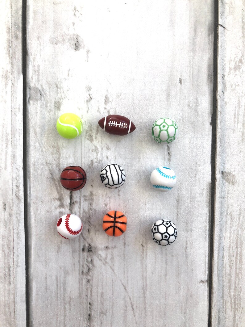 K&ouml;nnte beinhalten: Neun Miniatur-Sportb&auml;lle, darunter ein Tennisball, ein American Football, ein Fu&szlig;ball, ein Basketball, ein Volleyball, ein Baseball und zwei weitere Fu&szlig;b&auml;lle. Die B&auml;lle sind wei&szlig; mit verschiedenen farbigen Details und in drei Reihen auf einer wei&szlig;en Holzoberfl&auml;che angeordnet.