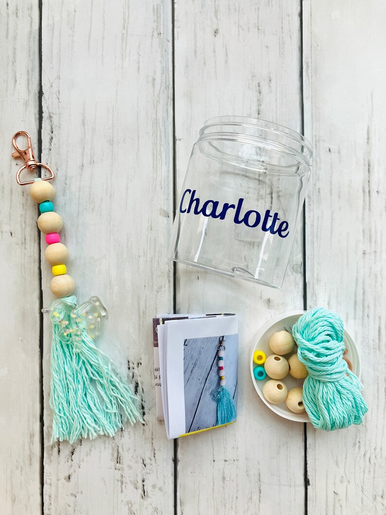 May include: A craft kit for making a beaded keychain with a turquoise tassel. The kit includes a clear plastic jar labeled "Charlotte", a small booklet with instructions, a skein of turquoise yarn, wooden beads, and colorful beads.