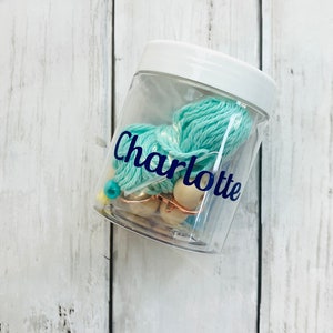 May include: A clear plastic jar with a white lid, filled with teal yarn and wooden beads. The jar has a blue label with the name "Charlotte" on it.
