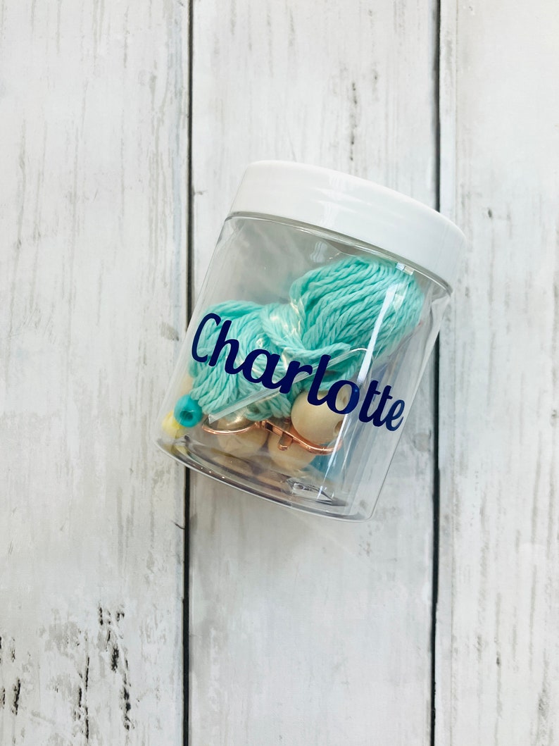 May include: A clear plastic jar with a white lid, filled with teal yarn and wooden beads. The jar has a blue label with the name "Charlotte" on it.