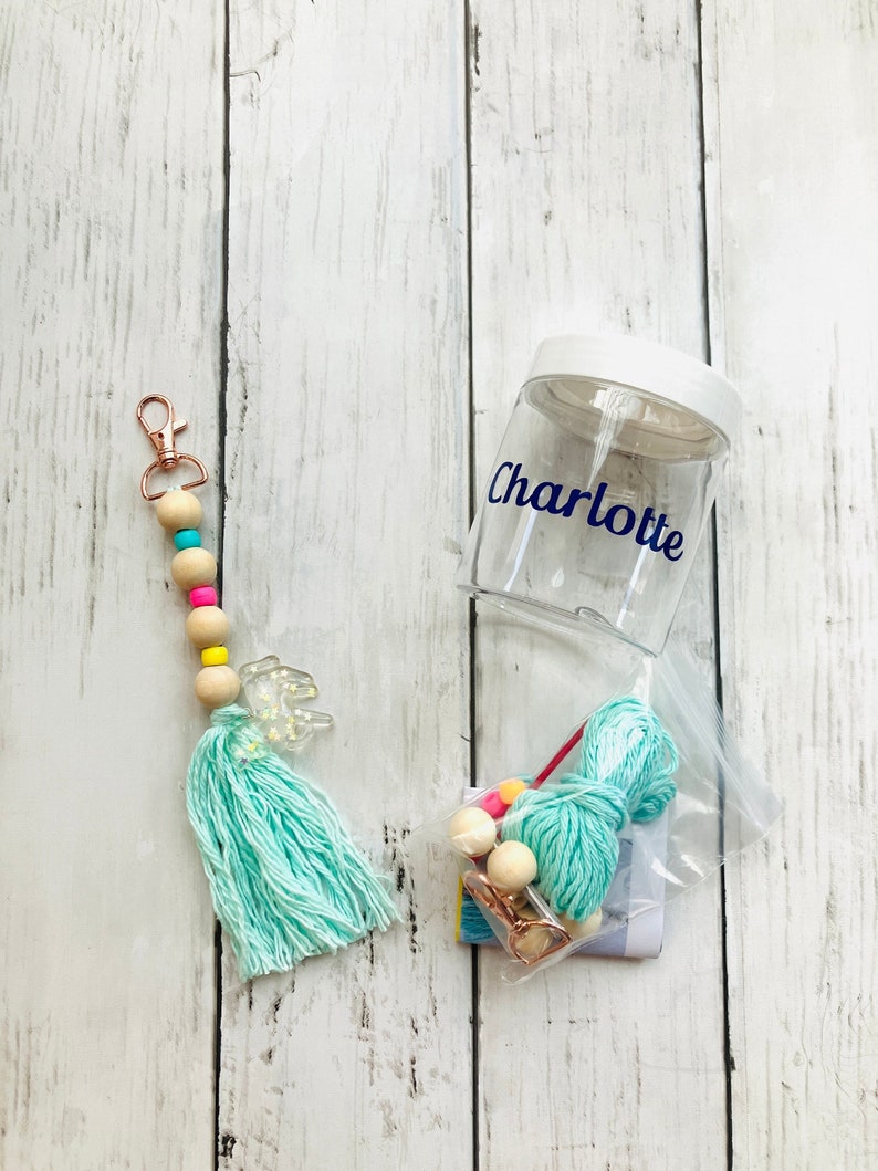 May include: A clear plastic jar with a white lid and the name "Charlotte" written on it. The jar is filled with colorful beads, yarn, and a small metal clip. A turquoise tassel keychain with wooden beads and a clear plastic charm hangs on the left side of the image.