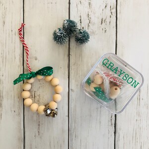 May include: A wooden bead wreath ornament with a green and white polka dot ribbon and a silver bell. The ornament is in a clear plastic container with the name "GRAYSON" written on it. There are also three small green and white pom-poms in the image.